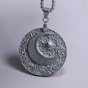 Könnte beinhalten: Ein runder, silberfarbener Metallanhänger mit einem erhabenen Halbmond- und Sonnendesign in der Mitte, umgeben von einem komplizierten, geprägten Rand aus wirbelnden Mustern. Der Anhänger hängt an einer silberfarbenen Metall-Boxkette.