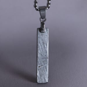 Puede incluir: Un colgante rectangular con una superficie metálica gris texturizada que presenta un patrón entrecruzado. El colgante está suspendido de una cadena de metal plateado mediante una pequeña anilla pulida, creando una pieza de joyería vertical.