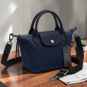May include: A navy blue handbag featuring a top flap with a silver-tone snap button, two short handles, and a detachable black shoulder strap with silver-tone metal clips. A rectangular tag labeled ENERGY hangs from the side of the bag.