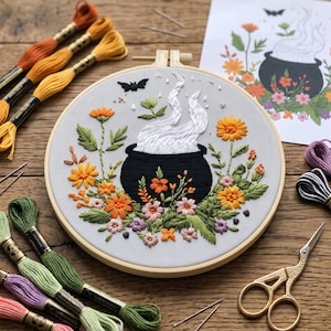 Peut inclure: Un tambour à broder circulaire en bois contenant un panneau en tissu avec un chaudron noir brodé dégageant de la vapeur blanche, entouré de fleurs orange, jaunes et roses avec des feuilles vertes et une petite chauve-souris noire.