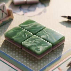 May include: Four rectangular mahjong tiles with a marbled green top surface and layered white and purple bases arranged in a square formation, accompanied by a tile rack displaying a tile with red Chinese characters and a black ink pen.