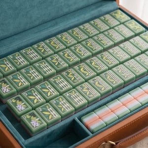 Pode incluir: Um conjunto de mahjong dentro de uma maleta de couro sint&eacute;tico castanho com interior em veludo azul-petr&oacute;leo. O conjunto inclui pe&ccedil;as retangulares verdes com ilustra&ccedil;&otilde;es de bambu e flores com marca&ccedil;&otilde;es num&eacute;ricas, al&eacute;m de uma fila de pe&ccedil;as pastel.