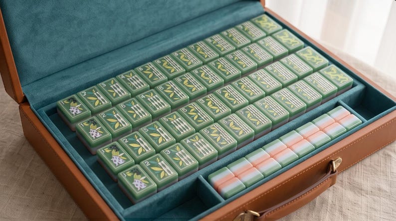 Pode incluir: Um conjunto de mahjong dentro de uma maleta de couro sint&eacute;tico castanho com interior em veludo azul-petr&oacute;leo. O conjunto inclui pe&ccedil;as retangulares verdes com ilustra&ccedil;&otilde;es de bambu e flores com marca&ccedil;&otilde;es num&eacute;ricas, al&eacute;m de uma fila de pe&ccedil;as pastel.