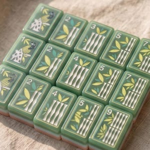 Pode incluir: Um conjunto de treze pe&ccedil;as retangulares verdes dispostas numa grelha. Cada pe&ccedil;a apresenta um acabamento brilhante com n&uacute;meros impressos e ilustra&ccedil;&otilde;es de caules de bambu ou p&aacute;ssaros estampados. As pe&ccedil;as t&ecirc;m uma base branca.