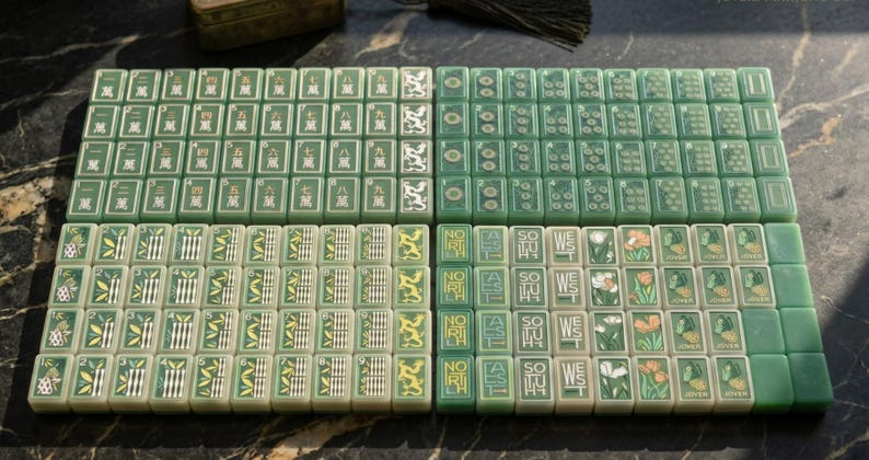 Pode incluir: Um conjunto completo de pe&ccedil;as retangulares de mahjong em tons de verde claro e escuro. As pe&ccedil;as apresentam caracteres chineses gravados, padr&otilde;es de bambu, desenhos florais e texto direcional incluindo North, East, South e West em branco.