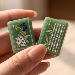 Pode incluir: Duas pe&ccedil;as retangulares verdes de mahjong com desenhos gravados. A pe&ccedil;a da esquerda exibe o n&uacute;mero 1 com um motivo de p&aacute;ssaro e folhagem. A pe&ccedil;a da direita exibe o n&uacute;mero 4 com um padr&atilde;o de bambu. Ambas possuem detalhes em tom dourado.