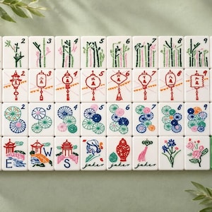 May include: A set of white Mahjong tiles arranged in four rows. The tiles feature colorful illustrations of bamboo stalks, lanterns, floral patterns, pagodas, and dragons. Some tiles include the letters N, E, W, S, and the word joker in script.