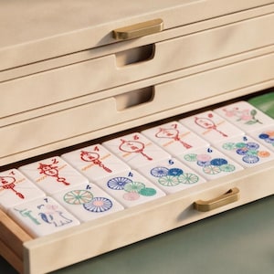 May include: A light-colored wooden storage chest with three drawers, featuring brass-toned corner accents and handles. The bottom drawer is open, revealing a set of white rectangular tiles with colorful graphic illustrations and numbers printed on them.
