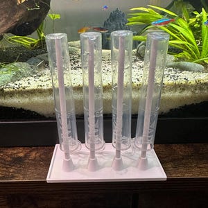 May include: A white plastic aquarium test tube holder with four clear tubes. Each tube has a pink rod inside. The tubes are in front of an aquarium with fish and plants.