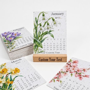 May include: A 2027 desk calendar set featuring floral illustrations for January, February, and March. The January card sits in a light wood stand with the text Custom Your Text. Each card displays a month, dates, and Your Custom Text.