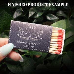 May include: A rectangular black matchbox featuring a white line art illustration of two swans facing each other. The text reads "Olivia & James" and "08/25/2027". The box contains wooden matches with red tips, and the top text reads "FINISHED PRODUCT EXAMPLE".