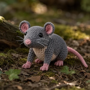 May include: A handmade crochet mouse in a natural outdoor setting. The gray mouse has pink accents on its ears, nose, feet, and tail. It has black eyes and white whiskers. The mouse is sitting on a bed of moss and leaves.