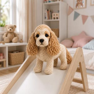 Op de afbeelding: Een gehaakt knuffeldier in de vorm van een spaniël met lichtbruine vacht, lange golvende oren en donkere ogen. Het pluche speelgoed staat op een lichtgekleurde houten glijbaan. De hond heeft een lichaam van textuurwol en een kleine zwarte neus.