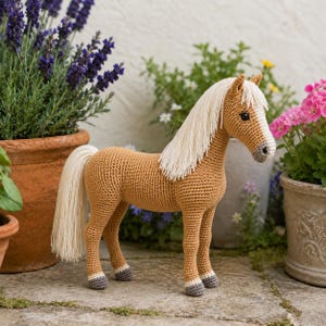 Könnte beinhalten: Gehäkeltes Pferd mit hellbraunem Körper, cremefarbener Mähne und Schweif. Die Hufe sind grau. Das Pferd steht auf einem Steinboden, umgeben von Topfpflanzen mit lila und rosa Blüten.