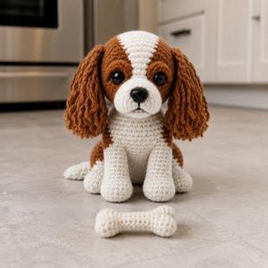 Puede incluir: Un perro Cavalier King Charles Spaniel tejido a ganchillo con marcas marrones y blancas, ojos negros grandes y orejas caídas. Un pequeño hueso tejido a ganchillo está delante del perro. El perro está sentado sobre una superficie gris claro.