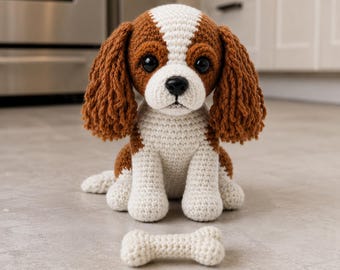 May include: A crocheted Cavalier King Charles Spaniel dog with brown and white markings, large black eyes, and floppy ears. A small, crocheted bone lies in front of the dog. The dog is sitting on a light gray surface.