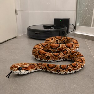 Könnte beinhalten: Eine gehäkelte Schlange in Braun-, Beige- und Schwarztönen, die einer Kornnatter ähnelt. Das handgefertigte Reptil weist ein detailliertes Muster, schwarze Augen und eine gespaltene Zunge auf. Die Schlange ist auf einem grauen Fliesenboden aufgerollt.