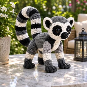 May include: A crocheted lemur figurine featuring a long, curved tail with alternating black, white, and gray stripes. The body is crafted from gray and white yarn, with large dark eyes, a black nose, and distinct black patches around the eyes.
