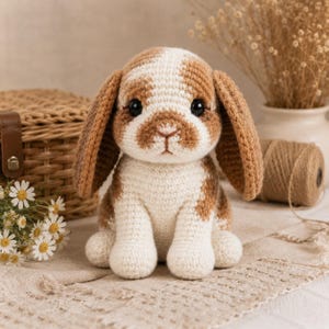 Könnte beinhalten: Ein handgehäkelter Hase mit braunen und weißen Flecken. Der Hase hat lange, schlaffe Ohren und große schwarze Augen. Er sitzt aufrecht auf einer cremefarbenen Oberfläche. Der Hase ist ein weiches Spielzeug, perfekt für ein Kinderzimmer oder als Dekorationsartikel.