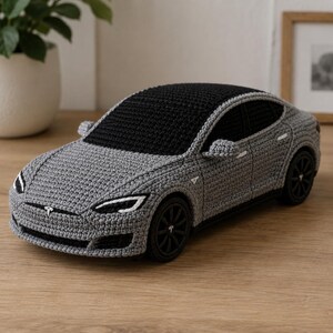 May include: Crocheted grey car with a black roof and black wheels. The car features white headlights and a Tesla logo. The handmade car is sitting on a wooden surface. The car is a model of a Tesla.