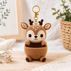 May include: A crocheted deer keychain with a gold-colored clasp. The deer has a brown and cream color scheme with small antlers, large eyes, and a small basket-like pouch. The keychain is sitting on a wooden surface.