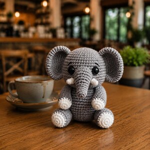 May include: A gray crocheted elephant stuffed animal with large ears, black plastic eyes, small white tusks, and white accents on the tips of its four feet. The toy is crafted with a visible yarn stitch pattern and sits in an upright position.