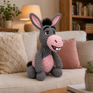 May include: A handmade crochet donkey plush toy in gray, pink, and black. The donkey has large ears with pink insides, a pink muzzle, and a smiling mouth. The plush is sitting on a beige couch with pillows.