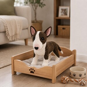 May include: A crocheted bull terrier soft toy made from dark brown and white yarn, standing in a light wood pet bed featuring a paw print cutout. Beside the bed sits a beige ceramic bowl with black paw prints and a twisted rope toy with brown and white fibres.