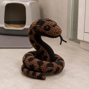May include: A handmade crocheted snake toy, featuring a pattern of brown and black. The snake is coiled, with its head raised and tongue extended. The toy has black eyes and a detailed, textured appearance.