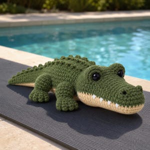 Puede incluir: Un juguete de cocodrilo verde hecho a mano con ganchillo, con vientre beige y dientes blancos. El cocodrilo tiene grandes ojos negros y está tumbado sobre una superficie gris oscura. El fondo muestra una piscina.