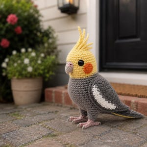 Peut inclure: Un perroquet cacatoès crocheté à la main, aux tons de gris, de jaune et d'orange. L'oiseau présente un design détaillé avec une crête, une tache orange sur la joue et des yeux noirs. L'oiseau est posé sur une surface en briques.