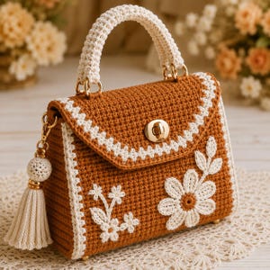 May include: A structured handbag crafted from brown crochet yarn featuring a cream-colored crocheted handle and trim. The front flap displays cream floral embroidery, a gold-tone metal turn-lock closure, and a matching cream tassel charm.