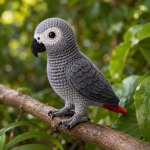 Peut inclure: Un perroquet crocheté à la main, dans des tons de gris, perché sur une branche brune. Le perroquet a un bec noir, des pieds noirs et une petite queue rouge. Le fond est un flou de feuillage vert.