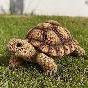 Op de afbeelding: Een gehaakte schildpadfiguur met een gedetailleerd, getextureerd schild in bruin- en beigetinten. De schildpad heeft een lichtbruine kop, poten en voeten, met zwarte ogen. Het rust op een bed van groen gras.