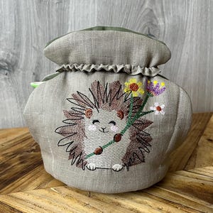 May include: A beige tea cosy featuring an embroidered hedgehog holding a flower branch. The hedgehog has brown quills and a smiling face. The cosy has a ruffled top and a green lid.