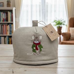 May include: A beige linen tea cozy featuring an embroidered brown mouse wearing a green jacket and a red scarf. A brown paper tag is attached to the top loop with a string, displaying a line drawing of a teapot and the text craftyorth.etsy.com.