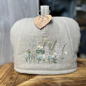 Puede incluir: Un calentador de té beige de lino con bordados de flores silvestres en blanco, azul y morado. Tiene un asa de bucle y una etiqueta que dice "Hand made with love".