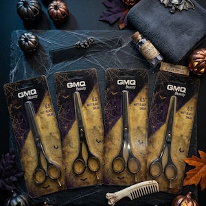 Puede incluir: Cuatro tijeras GMQ Beauty de 6.5 pulgadas en negro mate en envases individuales. El empaque presenta un diseño de telaraña y murciélagos. El set incluye dos tijeras de entresacar y dos tijeras de corte, todas con mangos y hojas negras.