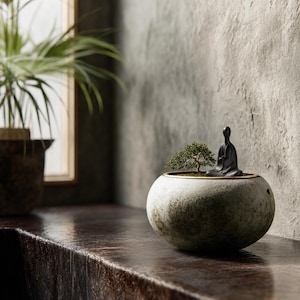 以下が含まれることがあります： 金色の縁が付いた、斑点のある丸い石のような鉢植えの小さな盆栽。 小さな黒い瞑想する人物が木の隣に座っています。 鉢は暗い木製の表面にあり、背景には窓と植物があります。