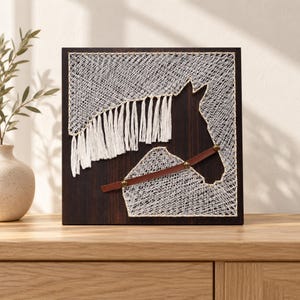 May include: A square dark wood wall art piece featuring a horse head silhouette created with white string art. The design includes a white yarn fringe mane and a brown leather strap across the horse's neck, secured with small brass-colored metal studs.