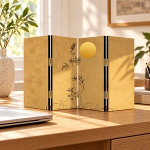 May include: A four-panel tabletop folding screen with a gold-toned finish. The screen features a black ink illustration of bamboo stalks and a solid yellow circle representing the sun. The panels are connected by small gold-coloured metal hinges.