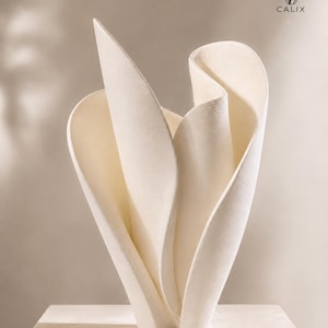 Può includere: Un oggetto scultoreo color crema con curve lisce e fluide a forma di petalo che si innalzano verso l'alto. Il pezzo presenta una finitura opaca e forme organiche ondulate. Nell'angolo in alto a destra, il testo CALIX appare sotto un'icona.