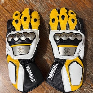 May include: A pair of black, white, and yellow motorcycle gloves featuring the Yamaha logo, metallic knuckle protection, and DTEC ENGINEERED text. The gloves have reinforced finger panels, a wrist strap, and protective padding on the palms and cuffs.