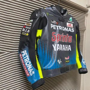 May include: A black, gray, and blue leather motorcycle jacket featuring Petronas, Yamaha, and Sprinta branding. The jacket includes neon yellow accents, sponsor logos such as Monster Energy and Akrapovic, and a gradient dot pattern on the front.