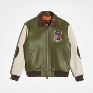 May include: Olive green leather jacket with cream-colored sleeves and brown accents. Features a brown collar, a zip-up front, and a patch on the chest with the text "AVIREX". The jacket has a ribbed hem and cuffs.