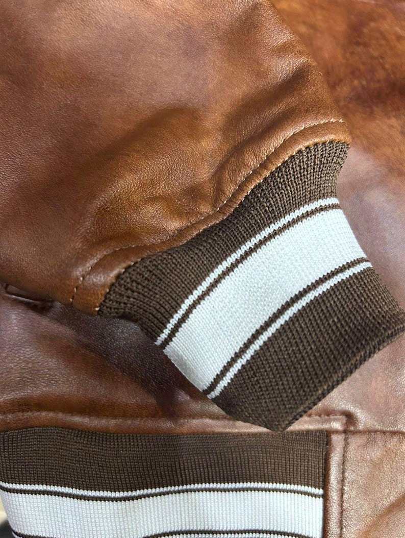Puede incluir: Primer plano de la manga de una chaqueta de cuero marr&oacute;n. El pu&ntilde;o es marr&oacute;n con dos rayas blancas. El cuero tiene un aspecto texturizado y un color marr&oacute;n rojizo c&aacute;lido. Las costuras son visibles a lo largo de las costuras.