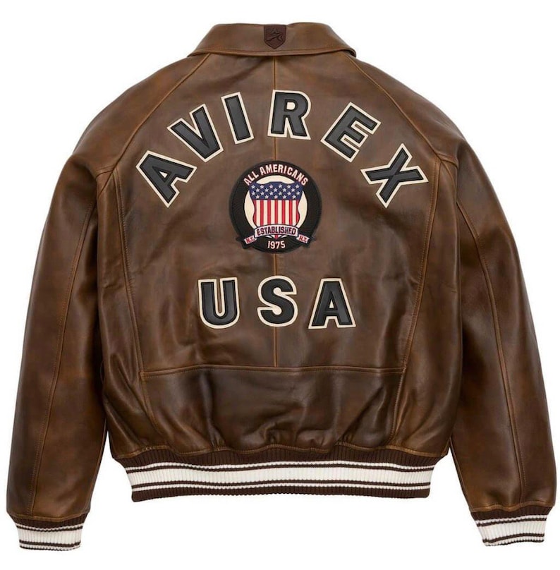 Puede incluir: Chaqueta de cuero marr&oacute;n con la palabra "AVIREX" en la espalda, junto con un parche que dice "ALL AMERICANS ESTABLISHED 1975 USA". La chaqueta tiene cuello y pu&ntilde;os acanalados con rayas blancas.