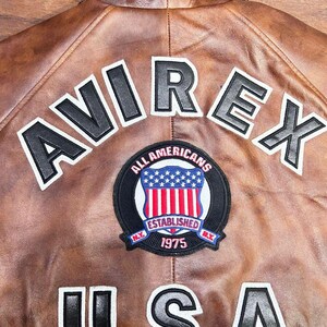 Puede incluir: Chaqueta de cuero marr&oacute;n con la palabra "AVIREX" en letras blancas y negras en la espalda. Un parche circular con un dise&ntilde;o de escudo y las palabras "ALL AMERICANS ESTABLISHED 1975" est&aacute; centrado debajo de las letras. La palabra "USA" est&aacute; debajo.