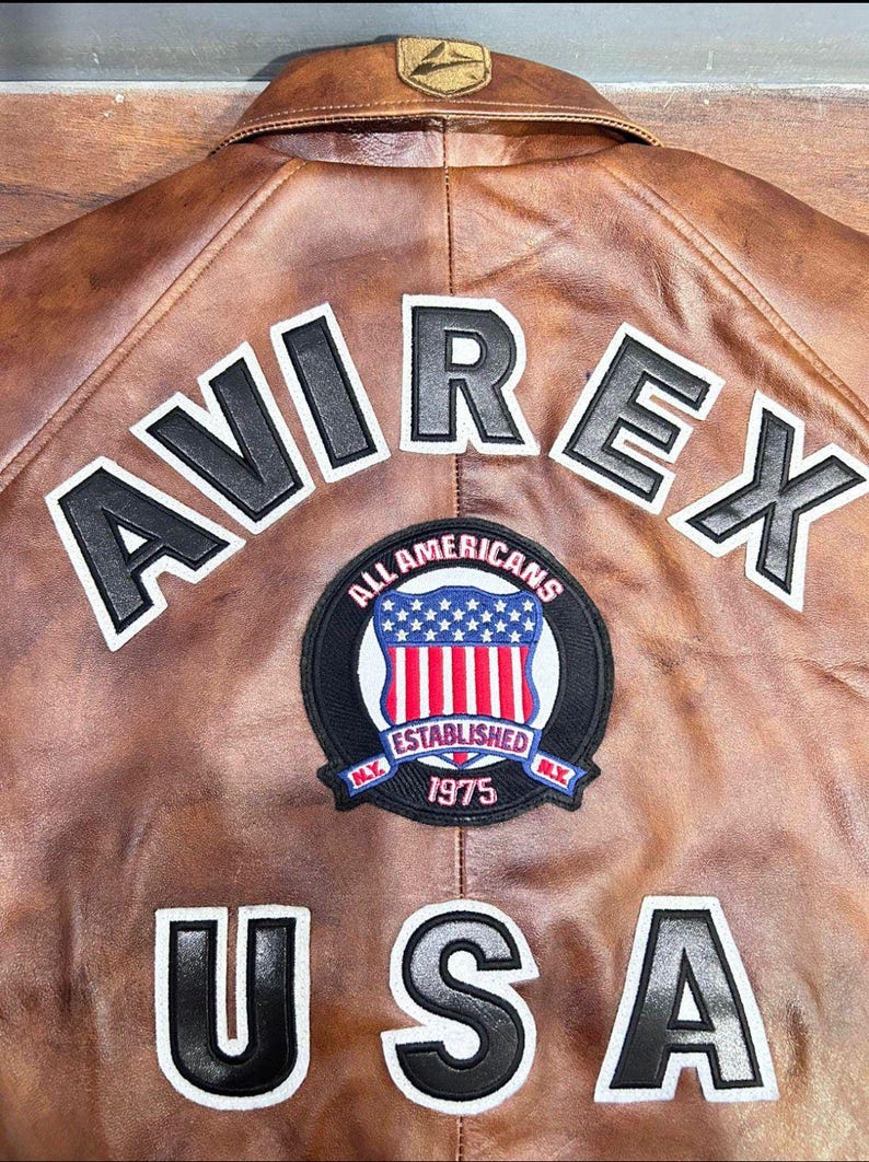 Puede incluir: Chaqueta de cuero marr&oacute;n con la palabra "AVIREX" en letras blancas y negras en la espalda. Un parche circular con un dise&ntilde;o de escudo y las palabras "ALL AMERICANS ESTABLISHED 1975" est&aacute; centrado debajo de las letras. La palabra "USA" est&aacute; debajo.