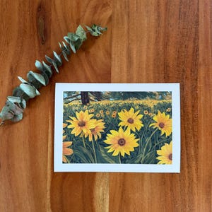 Puede incluir: Una impresión artística rectangular que representa un campo de girasoles amarillos con hojas verdes y un árbol al fondo. La impresión está sobre un fondo blanco y se coloca sobre una superficie de madera. Una ramita de eucalipto a la izquierda.